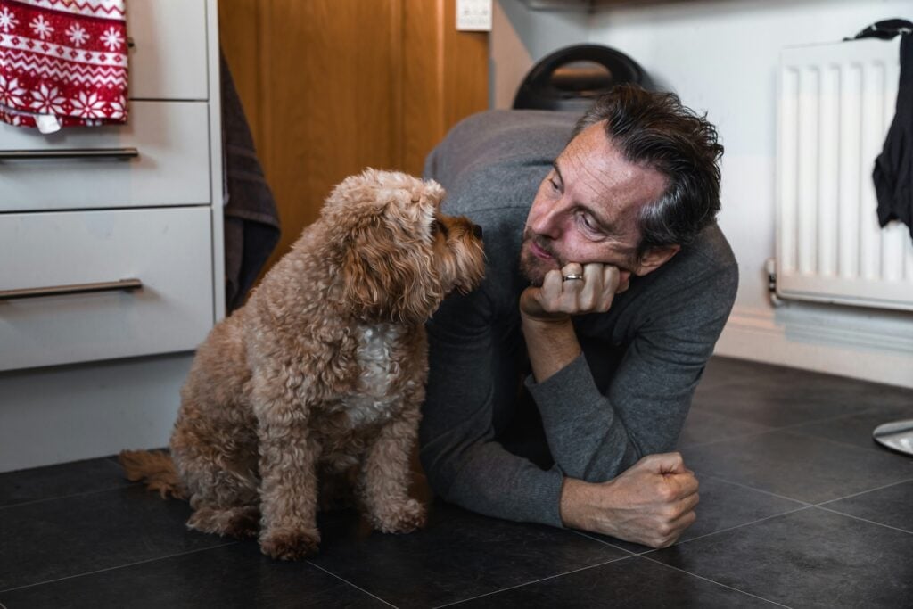 A man lies on a kitchen floor, propped on one arm, smiling and looking affectionately at a small, curly-haired dog sitting nearby—sharing the kind of quiet joy you might imagine before you get a Cavapoo.